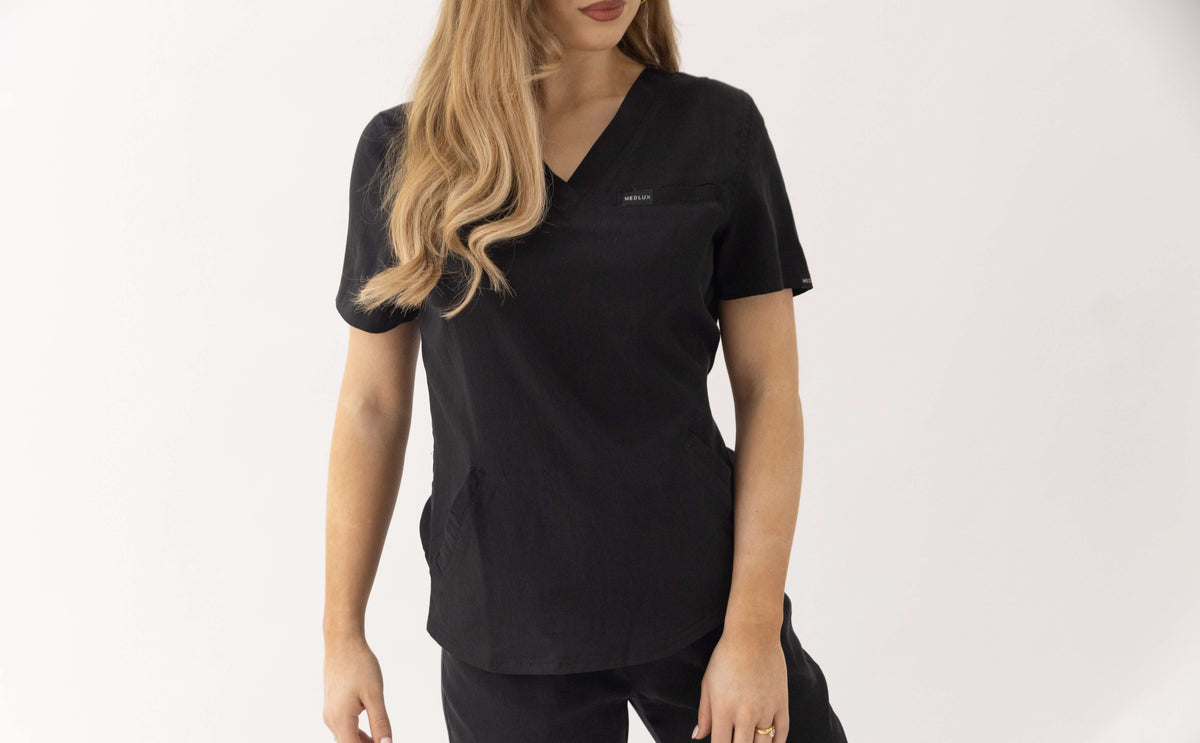 A woman wearing a black short-sleeve V-neck scrub top with a chest pocket, shown from the waist up against a plain light background.