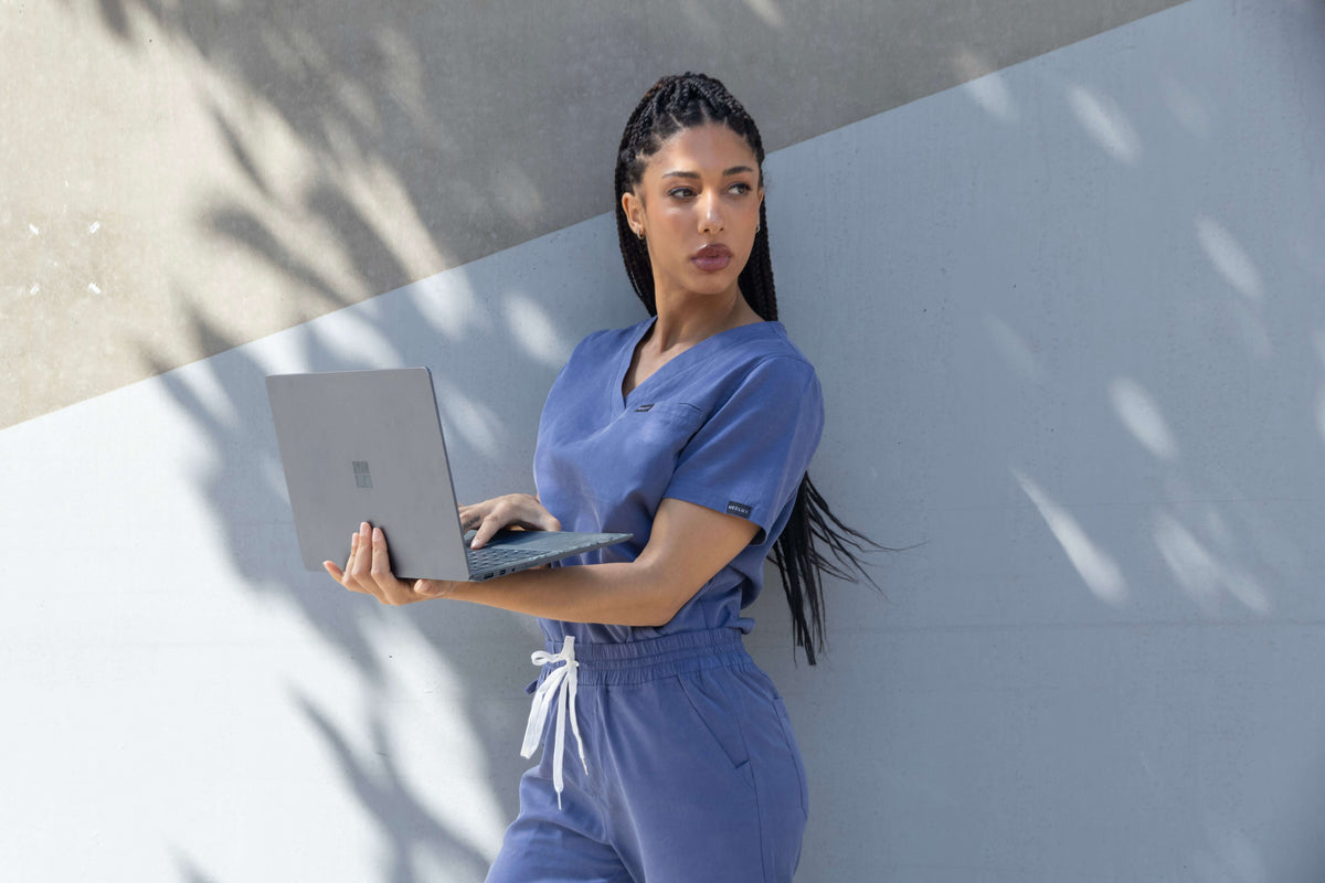 A woman wearing blue medical scrubs holds a laptop while leaning against a concrete wall with dappled shadows.