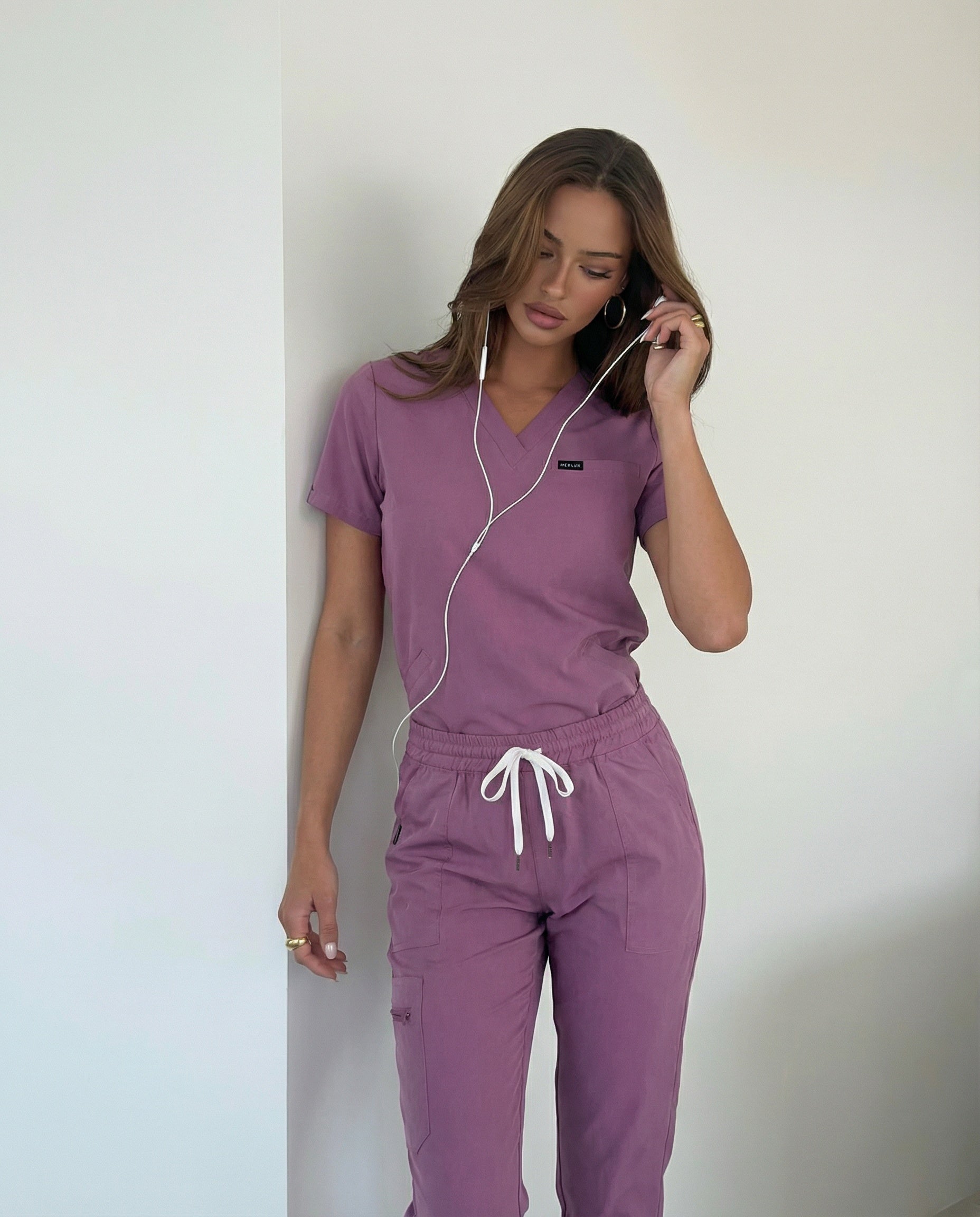 Hot girl wearing Australian purple scrub set with white earbuds against a plain background