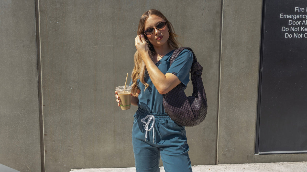 A woman wearing teal blue hospital scrubs and sunglasses stands against a grey concrete wall, holding an iced coffee and carrying a dark patterned shoulder bag.