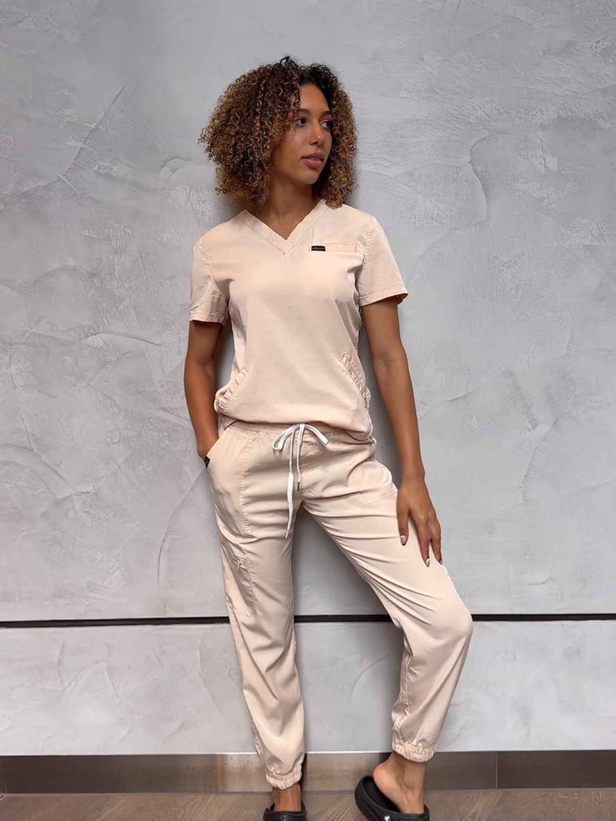 Nurse wearing cream stylish scrubs, leaning against grey wall
